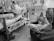 Two individuals are in a hospital room. One person is lying in a hospital bed with medical equipment around, engaging with a tray of food. The other person is sitting in a chair, using a smartphone, dressed in comfortable clothing.