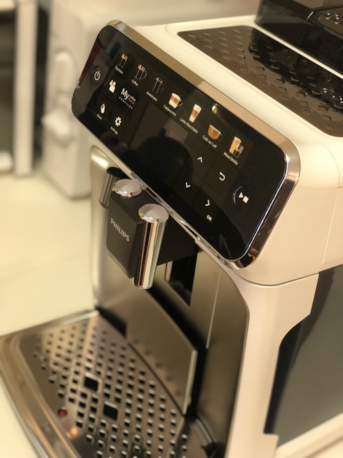 A sleek coffee machine with a user-friendly interface featuring options for various coffee types. The front panel is glossy with a digital display and several touch icons. The machine's metallic drip tray is visible at the bottom.