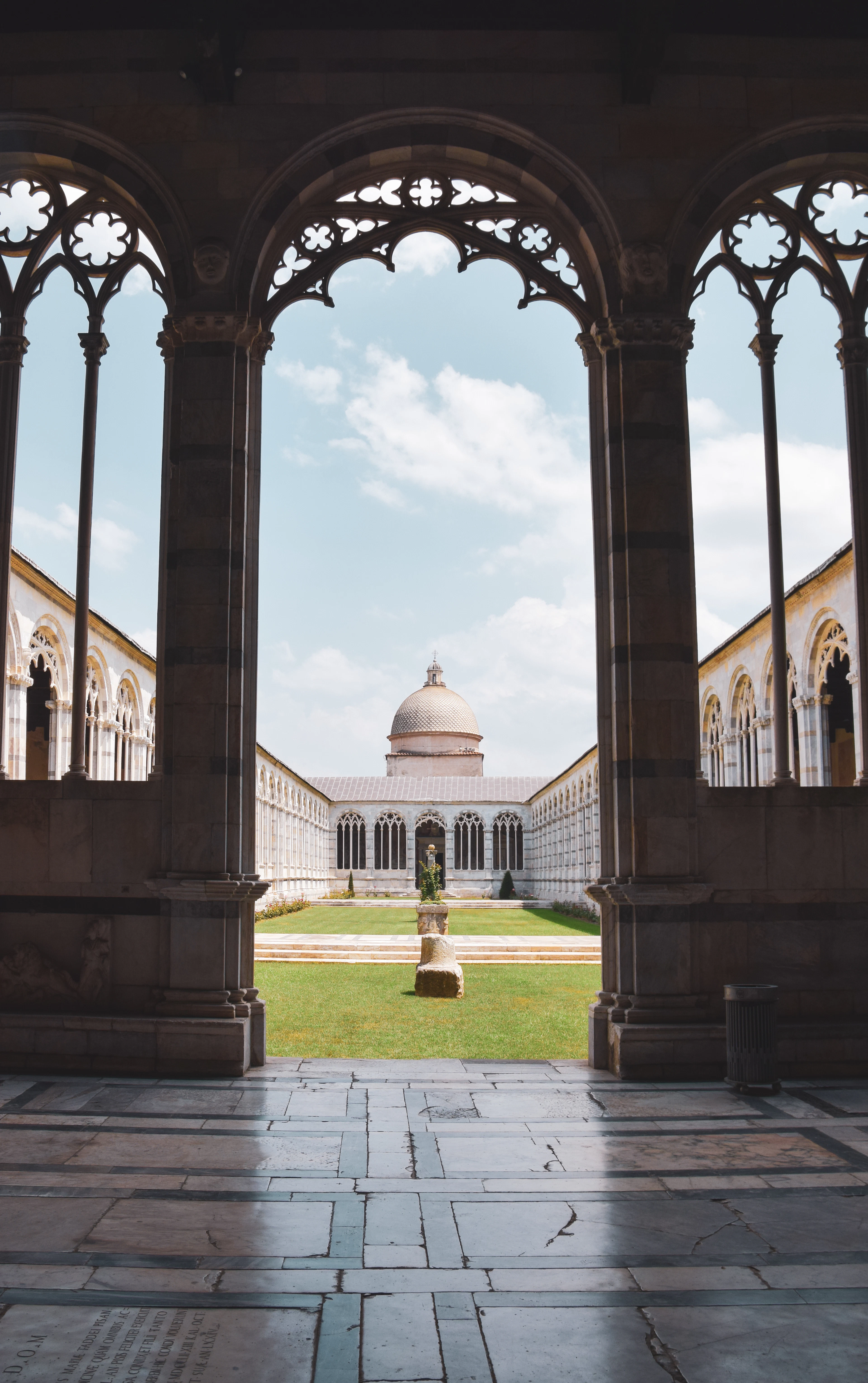 Inside the Camposanto Monumentale in Pisa, the delicate Gothic stone tracery of the cloister arcade framing the sunlit lawn of the inner courtyard and the small domed chapel on the far side.