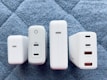 Four white charging adapters are arranged vertically on a quilted gray-blue surface. Each adapter differs slightly in design and size but commonly features USB ports, with some labeled for power delivery (PD) and specific wattages.