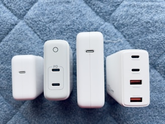 Four white charging adapters are arranged vertically on a quilted gray-blue surface. Each adapter differs slightly in design and size but commonly features USB ports, with some labeled for power delivery (PD) and specific wattages.