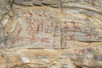 Ancient rock art featuring various human and animal figures painted with red pigment on a rocky surface. The figures appear to be engaged in activities, possibly depicting scenes from daily life or rituals.