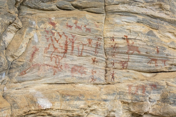 Ancient rock art featuring various human and animal figures painted with red pigment on a rocky surface. The figures appear to be engaged in activities, possibly depicting scenes from daily life or rituals.