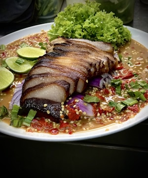 A plate of deliciously presented food features slices of grilled or roasted meat with a slightly charred exterior, served on a bed of colorful sauce. Fresh green lettuce, sliced purple onions, and multiple lime wedges complement the dish, adding vibrancy and contrasting textures.