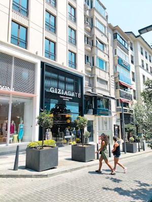 A busy urban street scene with shops and tall modern buildings. People are walking on the sidewalk past large windows displaying mannequins in fashionable clothing. Potted plants decorate the area, adding greenery to the urban environment.