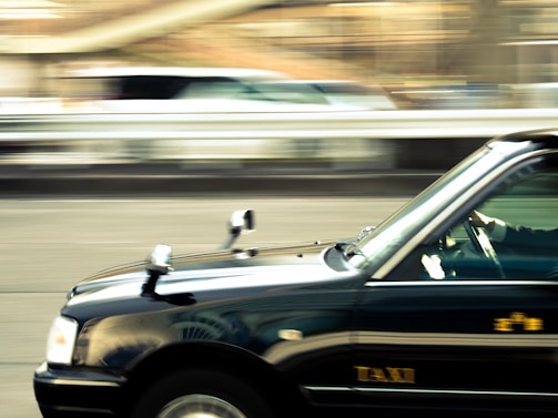 A black taxi is captured in motion with a blurred background, suggesting speed and movement. The focus is sharp on the taxi, with visible details like side mirrors and the word 'TAXI' on the door.