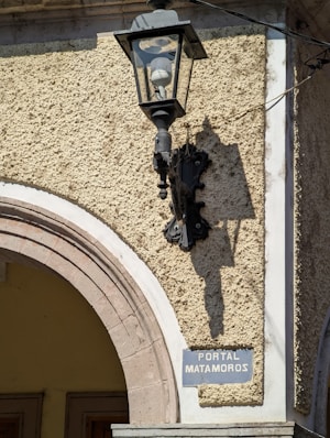 A vintage street lamp is mounted on a textured stone wall next to an arched doorway. The lamp casts a shadow on the wall, and a weathered sign reads 'Portal Matamoros' beneath it.