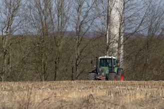 A green tractor is positioned in a harvested field with rows of stubble left behind. Behind the tractor, leafless trees form a dense backdrop, and a tall, cylindrical wind turbine tower can be partially seen through the trees.