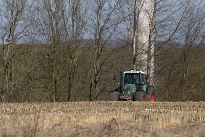 A green tractor is positioned in a harvested field with rows of stubble left behind. Behind the tractor, leafless trees form a dense backdrop, and a tall, cylindrical wind turbine tower can be partially seen through the trees.