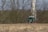 A green tractor is positioned in a harvested field with rows of stubble left behind. Behind the tractor, leafless trees form a dense backdrop, and a tall, cylindrical wind turbine tower can be partially seen through the trees.