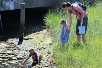 A woman with a backpack stands next to a young girl in a blue dress, and they are looking at a boy crouched near a small stream or pond. The boy is playing with water and mud. They are surrounded by tall green grass and there is a concrete structure in the background.
