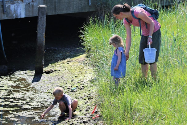 A woman with a backpack stands next to a young girl in a blue dress, and they are looking at a boy crouched near a small stream or pond. The boy is playing with water and mud. They are surrounded by tall green grass and there is a concrete structure in the background.