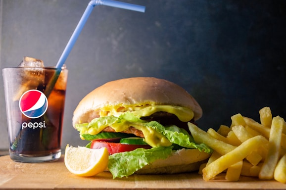 A delicious meal featuring a juicy burger with lettuce, cheese, and tomato, accompanied by a side of golden French fries. Next to the meal is a glass filled with Pepsi and ice, topped with a blue straw. A lemon wedge is also present in the foreground.