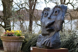 A stone sculpture with abstract facial features is displayed outdoors surrounded by foliage. The sculpture has exaggerated eyes and mouth, creating a whimsical expression. Nearby, a large pot filled with colorful flowers adds a pop of color to the scene. Leafless trees and a body of water create a peaceful, natural backdrop.
