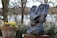 A stone sculpture with abstract facial features is displayed outdoors surrounded by foliage. The sculpture has exaggerated eyes and mouth, creating a whimsical expression. Nearby, a large pot filled with colorful flowers adds a pop of color to the scene. Leafless trees and a body of water create a peaceful, natural backdrop.