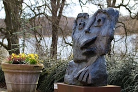 A stone sculpture with abstract facial features is displayed outdoors surrounded by foliage. The sculpture has exaggerated eyes and mouth, creating a whimsical expression. Nearby, a large pot filled with colorful flowers adds a pop of color to the scene. Leafless trees and a body of water create a peaceful, natural backdrop.