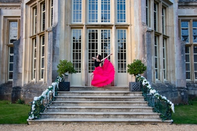 A person wearing a vibrant pink dress performs an elegant dance pose on the steps of a grand building. The architecture features large windows and detailed stonework. The steps are flanked by decorative greenery and white flower arrangements.