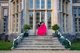 A person wearing a vibrant pink dress performs an elegant dance pose on the steps of a grand building. The architecture features large windows and detailed stonework. The steps are flanked by decorative greenery and white flower arrangements.
