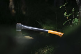 A single axe with an orange and black handle is embedded into a wooden log or stump. The scene is surrounded by greenery, suggesting a forest or wooded area. The lighting is dim, with sunlight partially illuminating the axe and the nearby foliage.