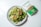 A fresh and colorful salad on a light gray plate includes a mix of greens, diced carrots, cucumbers, and tomatoes, topped with snap peas and a dried red pepper. Next to the plate, there's a package of freekeh with green and white labeling.