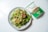A fresh and colorful salad on a light gray plate includes a mix of greens, diced carrots, cucumbers, and tomatoes, topped with snap peas and a dried red pepper. Next to the plate, there's a package of freekeh with green and white labeling.