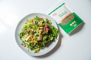 A fresh and colorful salad on a light gray plate includes a mix of greens, diced carrots, cucumbers, and tomatoes, topped with snap peas and a dried red pepper. Next to the plate, there's a package of freekeh with green and white labeling.