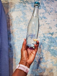 A hand is holding a clear glass bottle with a blue cap. The bottle features a label that reads 'Mayers Natural Spring Water.' The background consists of a textured surface with a mix of blue and beige tones.