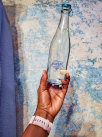 A hand is holding a clear glass bottle with a blue cap. The bottle features a label that reads 'Mayers Natural Spring Water.' The background consists of a textured surface with a mix of blue and beige tones.
