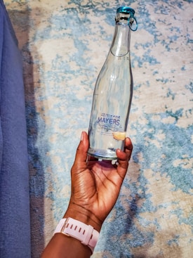A hand is holding a clear glass bottle with a blue cap. The bottle features a label that reads 'Mayers Natural Spring Water.' The background consists of a textured surface with a mix of blue and beige tones.