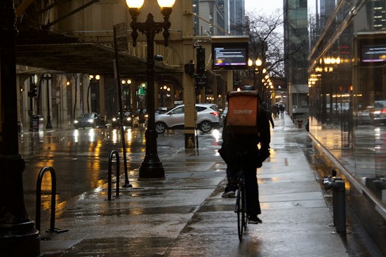 A cyclist with a delivery backpack rides on a wet city street lined with illuminated streetlights and reflected building lights, creating a moody urban atmosphere. Cars are visible in the background, and the street is flanked by tall buildings.