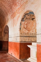 An ornate historical interior with intricate patterns and artwork on the walls and ceiling. The arches and borders are decorated with floral and geometric designs, primarily in earthy tones. There is a white architectural element resembling a mantle or altar piece on a red background wall.