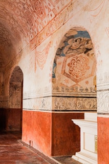 An ornate historical interior with intricate patterns and artwork on the walls and ceiling. The arches and borders are decorated with floral and geometric designs, primarily in earthy tones. There is a white architectural element resembling a mantle or altar piece on a red background wall.