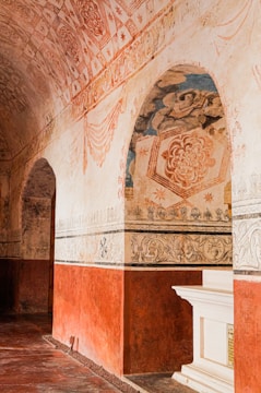 An ornate historical interior with intricate patterns and artwork on the walls and ceiling. The arches and borders are decorated with floral and geometric designs, primarily in earthy tones. There is a white architectural element resembling a mantle or altar piece on a red background wall.