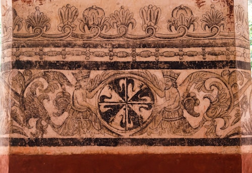 An intricately designed mural featuring ornate patterns and mythical figures. The artwork includes two crowned figures with wings, positioned symmetrically on either side of a circular, floral motif. Above them, a decorative border with floral and geometric elements adds complexity to the piece. The artwork is executed in a monochrome palette, set against a textured background.