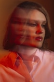 A person with a blurred double exposure effect, giving a sense of motion or introspection. The photograph features warm lighting with shades of red and orange overlaying a neutral background. The subject wears a light-colored shirt and appears contemplative.