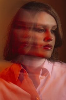 A person with a blurred double exposure effect, giving a sense of motion or introspection. The photograph features warm lighting with shades of red and orange overlaying a neutral background. The subject wears a light-colored shirt and appears contemplative.