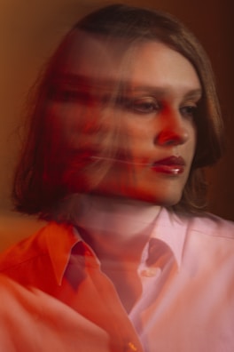A person with a blurred double exposure effect, giving a sense of motion or introspection. The photograph features warm lighting with shades of red and orange overlaying a neutral background. The subject wears a light-colored shirt and appears contemplative.