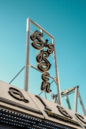 A vintage neon sign with the word 'Big' prominently displayed is mounted on a metallic framework against a clear blue sky. Below it, part of the structure appears with retro-styled elements.