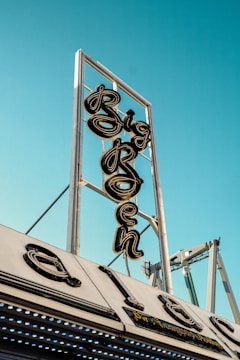 A vintage neon sign with the word 'Big' prominently displayed is mounted on a metallic framework against a clear blue sky. Below it, part of the structure appears with retro-styled elements.