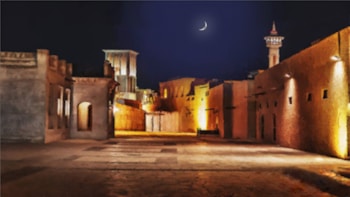 A dimly lit, historical street scene at night with traditional Middle Eastern architecture. The narrow street is flanked by old buildings with intricate designs and a visibly clear sky showcasing a crescent moon. Street lamps cast a soft yellow glow on the pathway, adding to the serene and atmospheric ambiance.