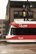 A red and white QLINE tram is stationary on a wet street against an urban backdrop of brick buildings. Overhead wires and streetlights are visible, adding to the city setting.