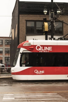 A red and white QLINE tram is stationary on a wet street against an urban backdrop of brick buildings. Overhead wires and streetlights are visible, adding to the city setting.