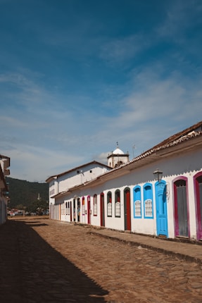 A cobblestone street is lined with colorful colonial-style buildings, each door painted in vibrant hues including blue, pink, and red. The architecture features white walls and arched window frames, and a small church tower is visible in the background under a clear blue sky.