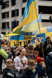 A group of people are gathered in a city street, holding and waving Ukrainian flags. They display signs with messages such as 'Stop the War' and 'Stand with Ukraine.' The scene is lively and crowded, with participants wearing various clothing, including traditional Ukrainian attire.