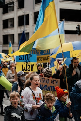 A group of people are gathered in a city street, holding and waving Ukrainian flags. They display signs with messages such as 'Stop the War' and 'Stand with Ukraine.' The scene is lively and crowded, with participants wearing various clothing, including traditional Ukrainian attire.