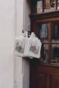 A set of canvas tote bags hang by a wooden door, displaying illustrations of a cityscape with the text 'Rothenburg'. The bags are attached to a metal hook and are priced at 3.50, as indicated by a small sign. The scene is softly lit, creating a cozy and quaint atmosphere.