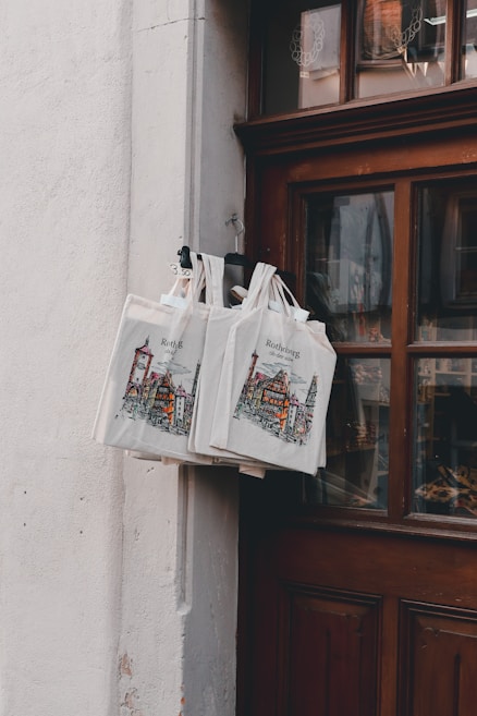 A set of canvas tote bags hang by a wooden door, displaying illustrations of a cityscape with the text 'Rothenburg'. The bags are attached to a metal hook and are priced at 3.50, as indicated by a small sign. The scene is softly lit, creating a cozy and quaint atmosphere.