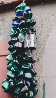 An outdoor lamp is creatively surrounded by a collection of colorful glass bottles, primarily in shades of green and blue. The bottles appear to be artistically arranged, hanging on a textured exterior wall.