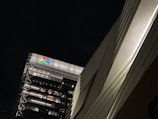 An urban scene featuring a tall building with modern architecture and illuminated windows. The brightly lit Google logo is prominently displayed on the top. The building is set against a dark night sky, and a nearby structure is visible on the right side with a textured surface.