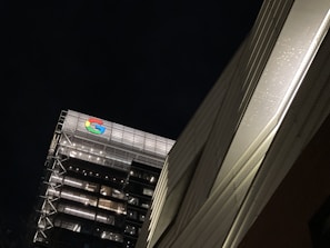 An urban scene featuring a tall building with modern architecture and illuminated windows. The brightly lit Google logo is prominently displayed on the top. The building is set against a dark night sky, and a nearby structure is visible on the right side with a textured surface.